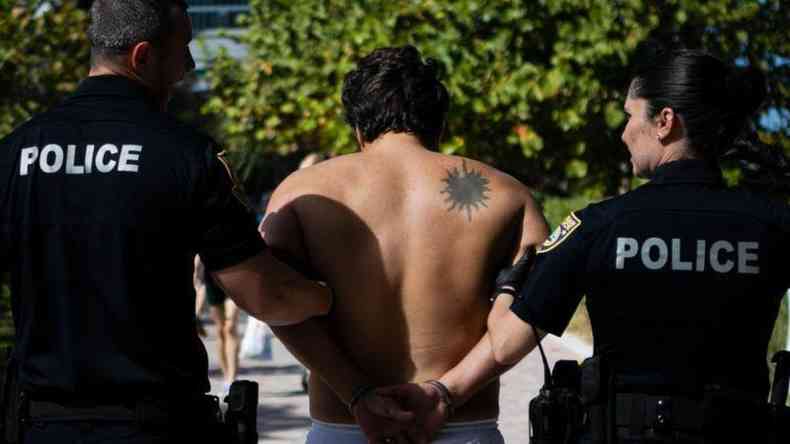 Homem sem camisa de costas sendo levado por dois policiais