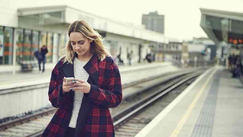 Mulher branca de cabelo loiro olhando para o celular em uma plataforma de trem