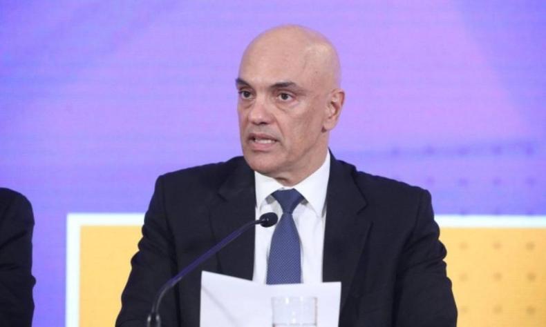 Alexandre de Moraes sentado segurando um papel enquanto fala em um microfone de mesa