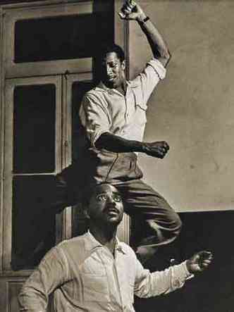 Fotografia captura cena de dois atores negros, vestidos com camisa branca. Enquanto um salta, outro olha para cima, com expresso de surpresa