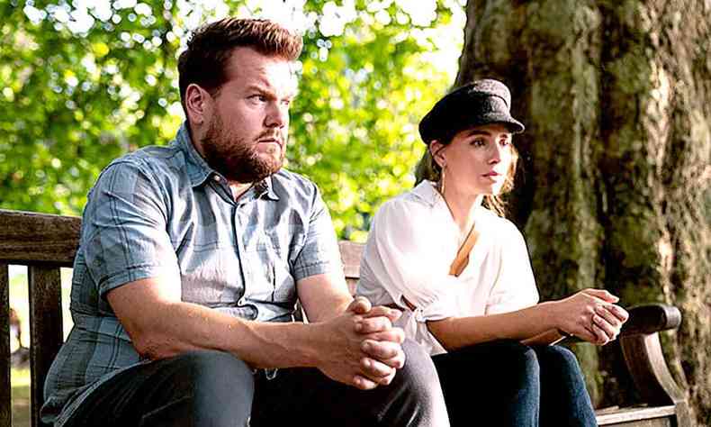 Os atores James Corden e Melia Kreiling, sentados em banco de madeira em parque, com expresso sria, em cena de 'vida de casal' 