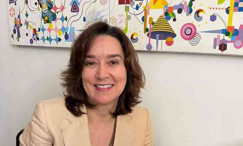 Maria José sorri pra a foto; ela está diante de um quadro com fundo branco e detalhes coloridos