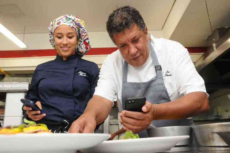 Olhando nos respectivos celulares, chefs Nayara Faria e Ivo Faria preparam prato em cozinha de restaurante