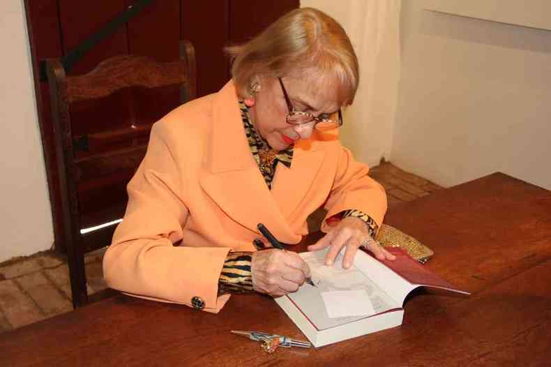 Vilma Guimares Rosa Reeves, escritora e filha de Guimares Rosa, veste um um blazer laranja. Ela est sentada em uma mesa autografando um de seus livros.
