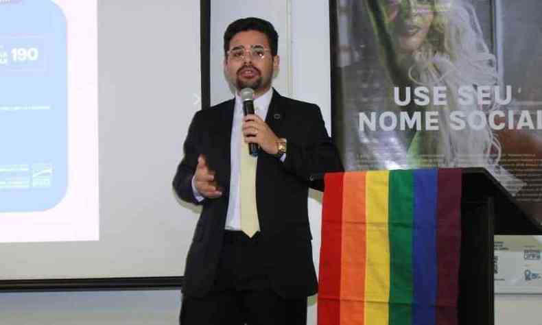 Gregory Rodrigues  um homem de cabelos e barba castanhos. Ele usa um terno preto com camisa branca e uma gravata preta. Ao seu lado, h um palanque com uma bandeira LGBT (com as cores do arco-ris) pendurada. Ele segura um microfone.
