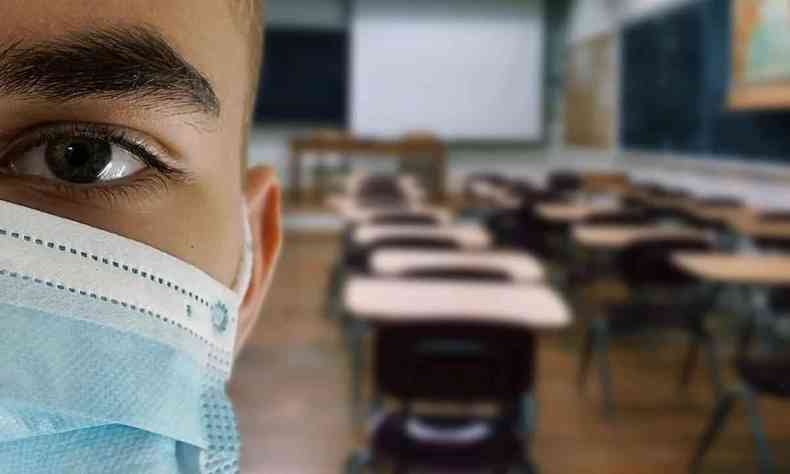 Imagem parcial do rosto de um jovem, usando mscara, dentro de uma sala de aula com carteiras vazias