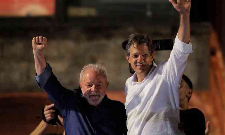Lula e Haddad, abraados com uma das mos para o alto. Lula esta a esquerda e veste uma camisa azul escuro e haddad veste uma camisa social branca