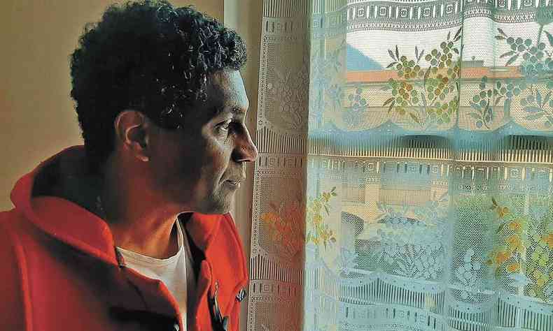 De camisa vermelha, fotografado de perfil, o escritor Edimilson de Almeida Pereira olha por uma janela coberta por cortina transparente