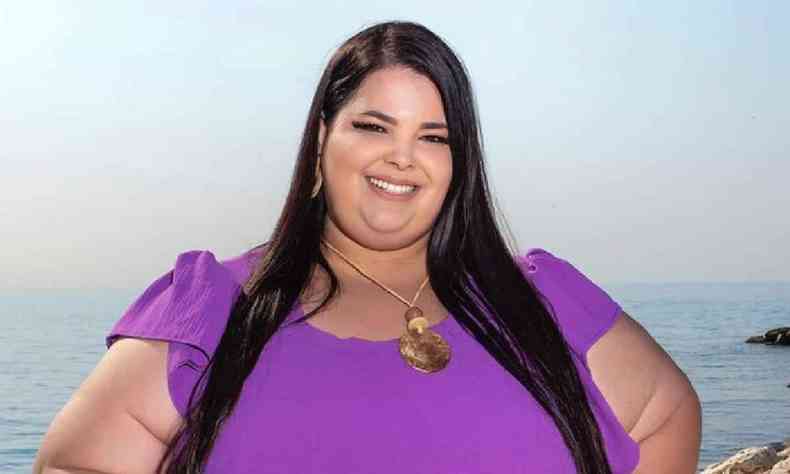 Juliana Nehme  uma mulher gorda, de cabelos castanhos escuros, longos e lisos, usa um vestido roxo com mangas curtas 