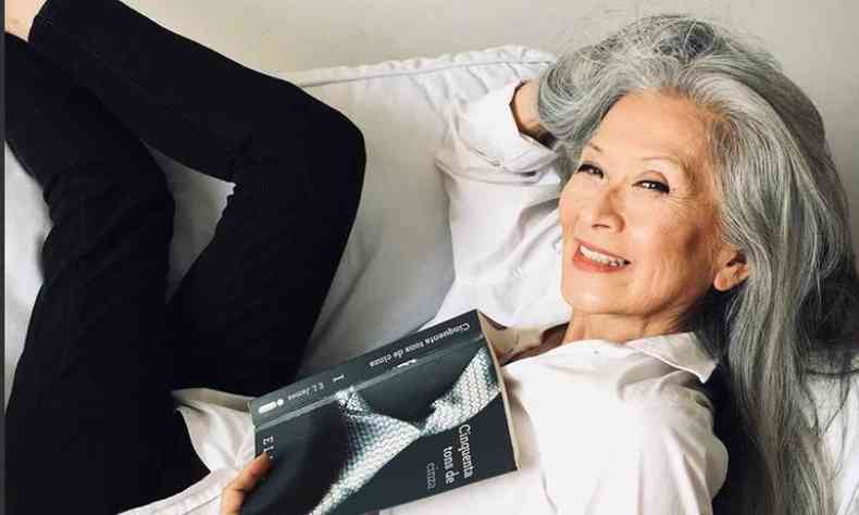 Rosa Saito, mulher de traços orientais deitada no sofá, usando camisa branca e calça preta, sorrindo para a câmera e segurando um livro