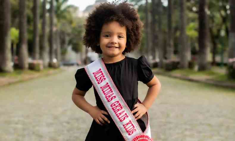 Duda, uma menina de 4 anos, cabelos black power, usando vestido preto, usa faixa de 'Miss Minas Gerais Kids 2022'