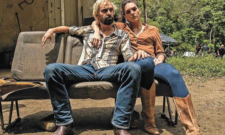 Atores Jlio Andrade e Julia Konrad, segurando revlver, esto abraados e sentados em sof no meio de um quintal em cena da srie 1 contra todos