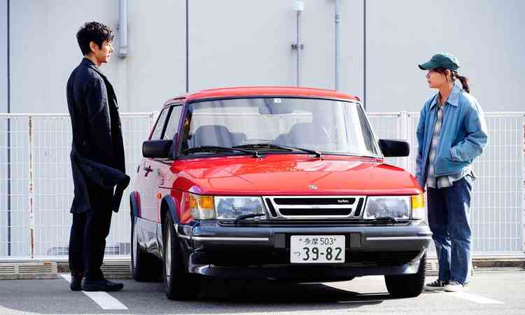 Homem e mulher orientais se olham e no meio deles j um carro vermelho, em cena do filme 'Drive my car'