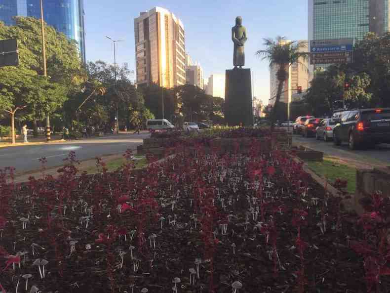 Praça Tiradentes em BH, canteiro de flores e cogumelos em primeiro plano, estátua Tiradentes no centro, carros parados no semáforo, árvores e prédios