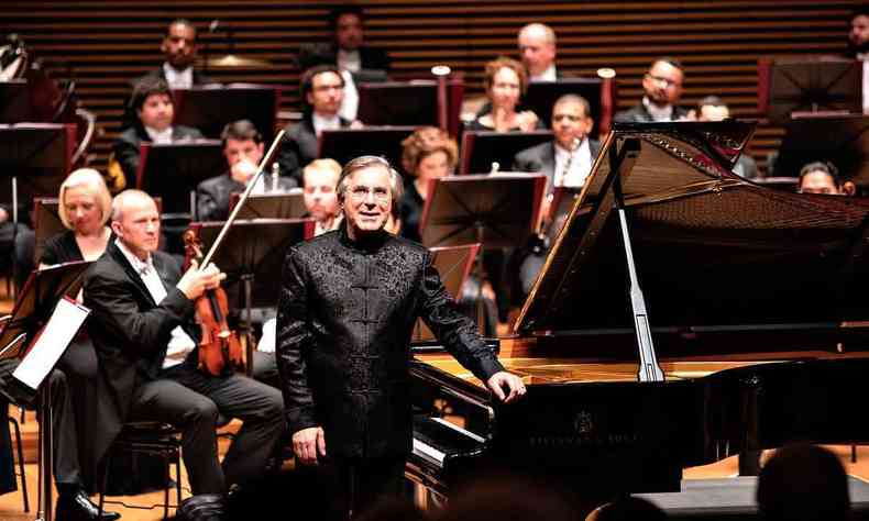 Arnaldo Cohen, de preto, de p, ao lado do piano, com msicos da Filarmnica de Minas Gerais atrs de si, no palco da Sala Minas Gerais