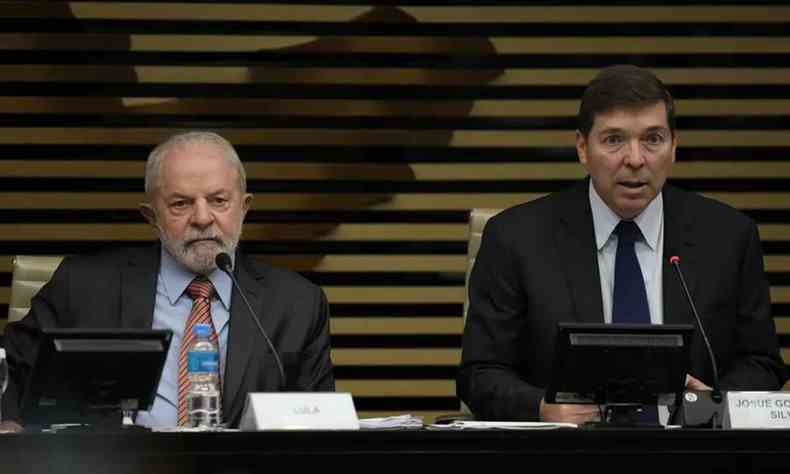 A imagem mostra Lula e Josue Gomes sentados um do lado do outro em uma bancada. Lula, homem branco de terno, sentado em frente a um microfone. Josue ao seu lado direito, homem branco de terno, tambm em frente a um microfone