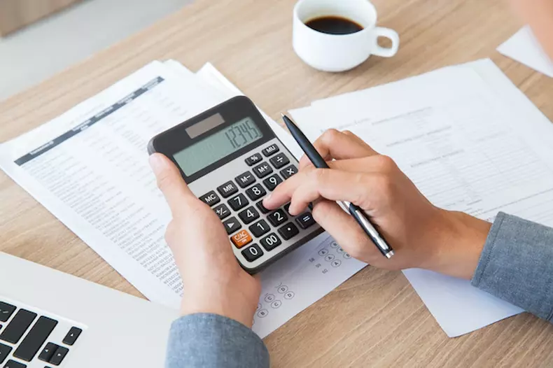 (foto: Freepik) mão segura calculadora sobre mesa cheia de papéis