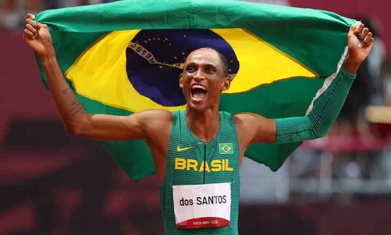 Alison  um homem negro de cabelos ralos e crespos. Ele sorri com a conquista da medalha de bronze nas Olimpadas de Tquio enquanto carrega a bandeira do Brasil sobre seu corpo.