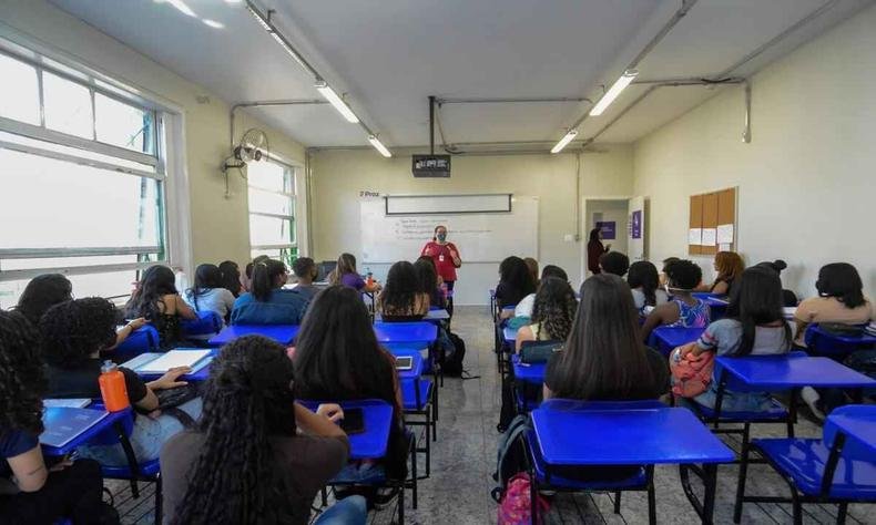 Crian�as em sala de aula com carteiras azuis e uma professora na frente de um quadro