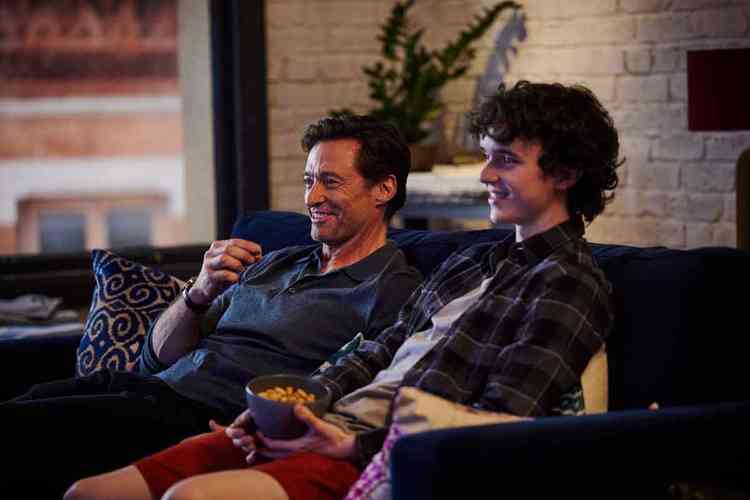 Os atores Hugh Jackman e Zen McGrath sorriem, sentados em sof, assistindo  TV, em cena de Um filho

