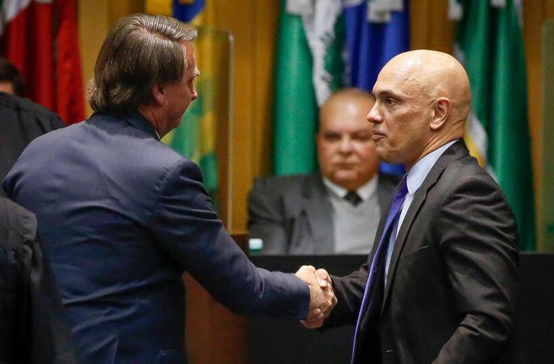 A açao também pede que o STF obrigue o governo a tomar medidas de prevenção para grupos vulneráveis, especialmente a comunidade LGBTQIA+(foto: SERGIO LIMA/AFP) Bolsonaro aperta a mão do ministro do STF Alexandre de Moraes. O fundo, há um homem careca de terno e gravata. O local é decorado com bandeiras do Brasil e dos estados brasileiros.