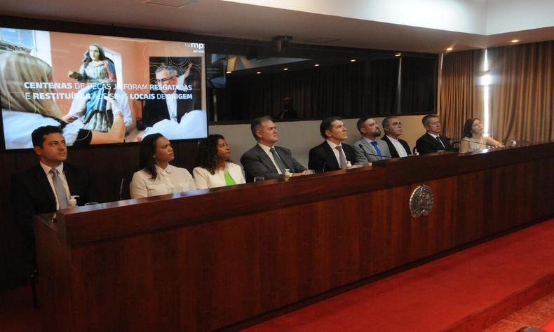 As autoridades do evento se encontram sentadas em uma mesa que se localiza em frente a um power point que apresenta o projeto Sondar. 