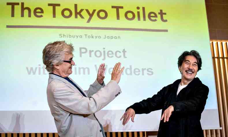 O cineasta Wim Wenders, de lado e sorrindo, ergue as mãos para o ator Koji Yakusho, que também sorri