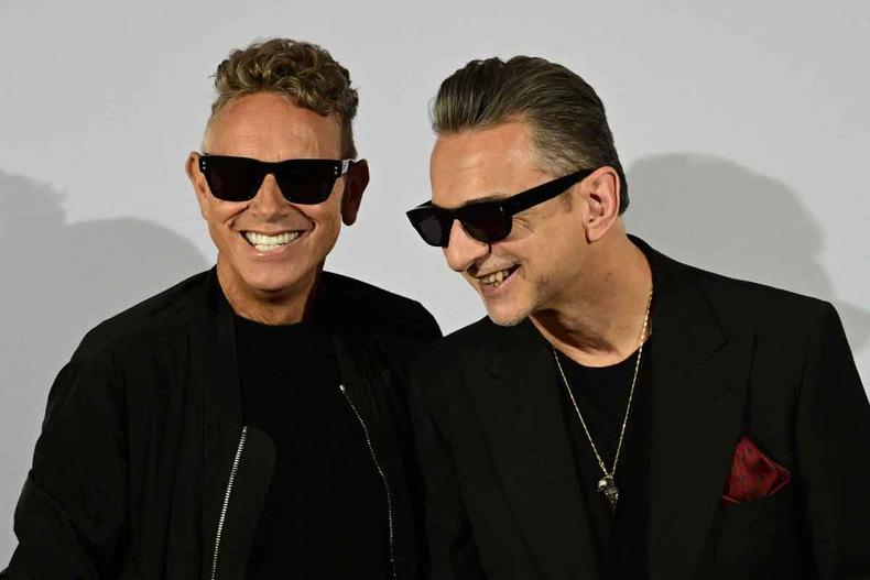 De casaco e culos pretos, Martin Gore e Dave Gahan posam para foto de p em frente a painel branco e sorriem 