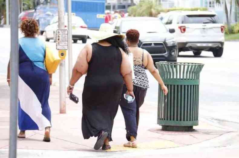 Mulher obesa, com vestido preto longo e chapu, caminha pela calada 