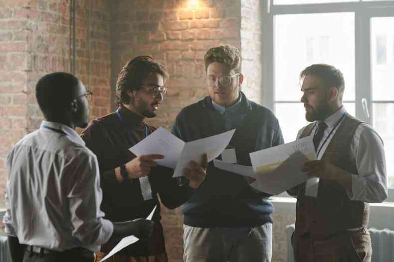 Homens em local de trabalho, todos de roupa social mostrando papis uns aos outros