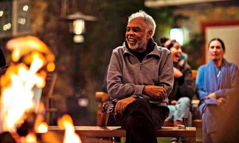 sentado num banco de madeira, vestindo calça preta e agasalho cinza, Gilberto Gil sorri 