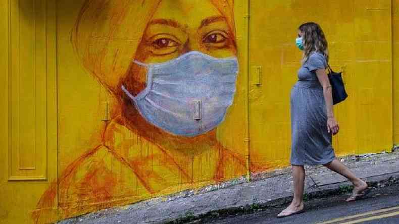 Gestante usando por máscara anda numa rua com um grafite de outra mulher, que também usa máscara