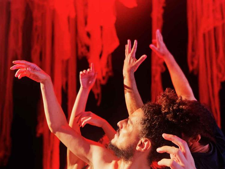 Bailarinos erguem os braços no palco, com fundo vermelho
