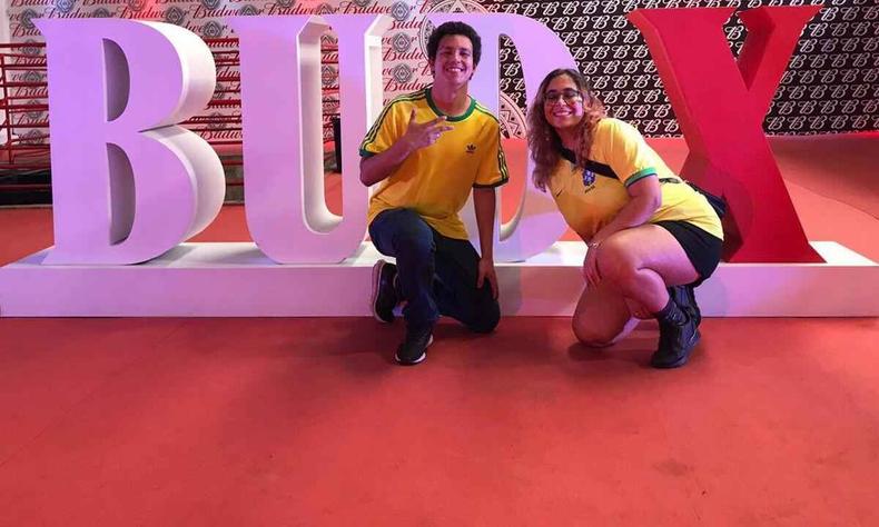 Thiago Abreu e Ana Carolina Fiuza, vestindo camisetas alusivas  Seleo Brasileira, sorriem e tm atrs de si instalao com enormes letras B, U, D e X nas cores lils e vermelha 