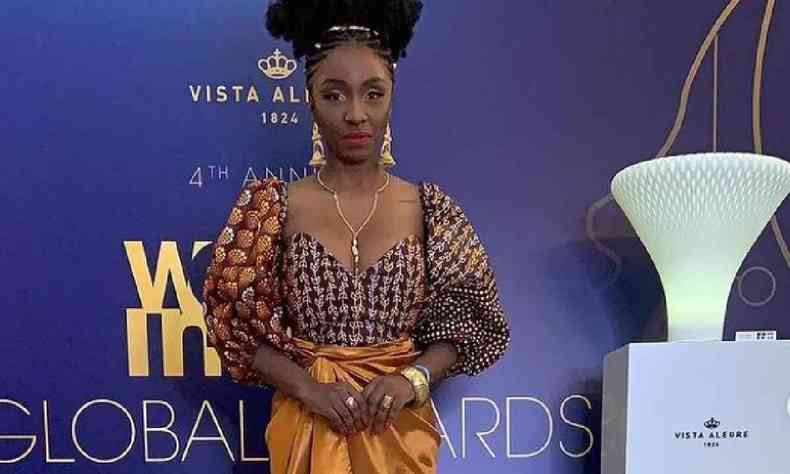 Nina Silva, mulher negra, com vestido de inspirao africana amarelo, posa diante de uma parede roxa com a logo do Women in Tech Global Awards