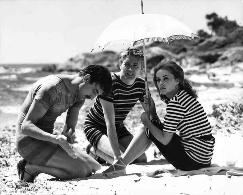 Os atores que interpretam o tringulo amoroso de Jules e Jim sentados na areia em cena do filme; Jeanne Moreau segura um guarda-sol