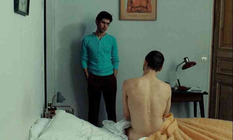 Homem conversa com outro homem nu, de costas, sentado na cama no filme Passages
