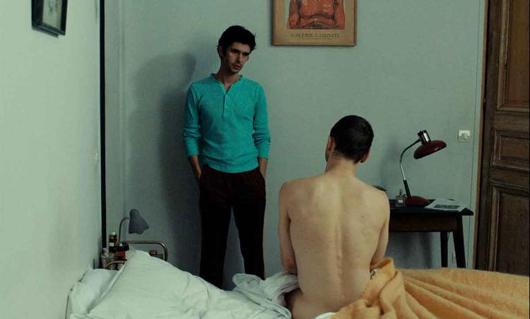 Homem conversa com outro homem nu, de costas, sentado na cama no filme Passages