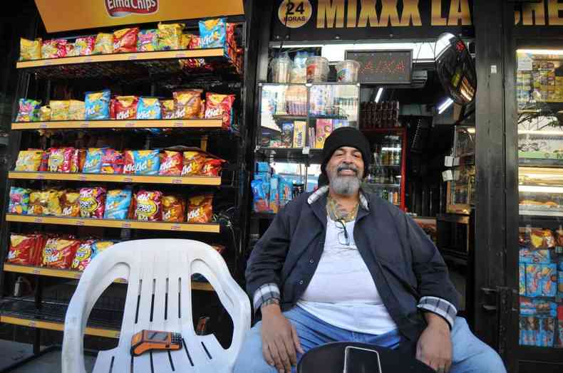 Edno Pereira est sentado em frente a uma loja; ele tem barba e cabelos grisalhos e usa um touca de frio