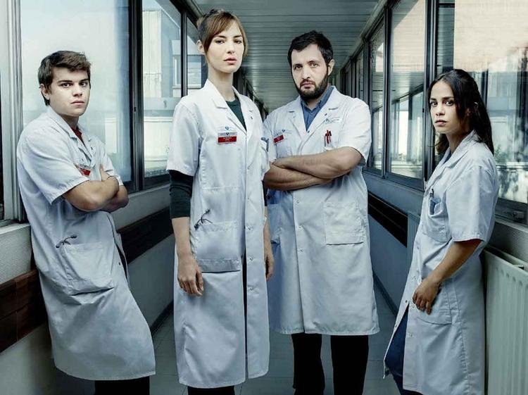 Atores com jaleco branco em corredor de hospital olham para a c�mera com express�o s�ria