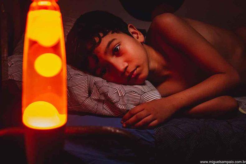 'Sideral', de Carlos Segundo, disputa uma vaga de curtas no Oscar(foto: Miguel Sampaio/Divulgação) Menino sem camisa deitado na cama olha para uma luminária laranja e amarelo em forma de foguete
