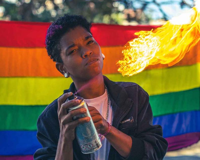 Grafiteiro Wanata segura lata de spray, de onde sai fogo, tendo ao fundo a bandeira com cores do arco-ris