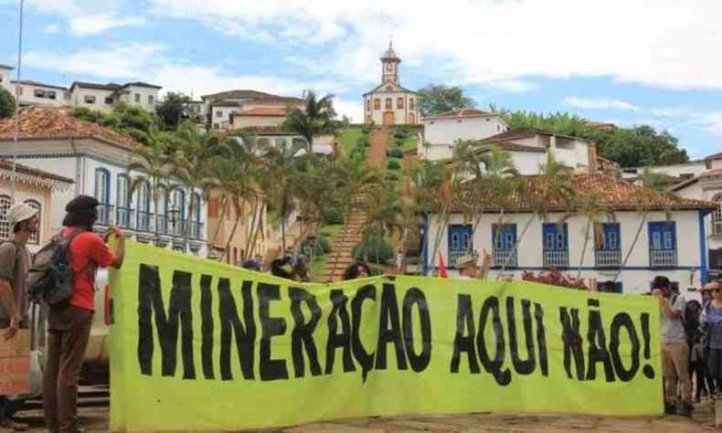 Pessoas em frente a uma praa segurando grande cartaz escrito 'minerao aqui no'