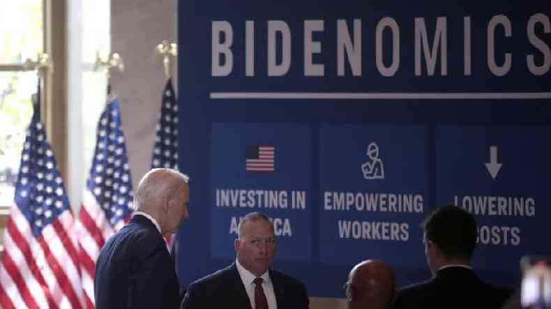 Biden e assessores perto de bandeiras dos EUA e painel com a palavra 'Bidenomics'