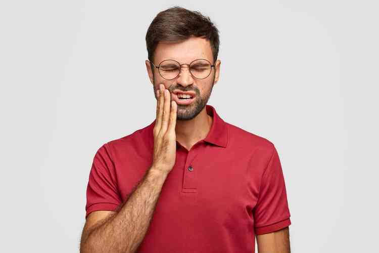 Homem branco com barba e camisa polo vermelha queixando-se de dor de dente 