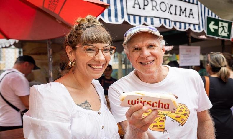 duas pessoas posando para a foto, uma mulher de camisa branca e um home tamb�m de camisa branca, mas que est� segurando uma salsicha