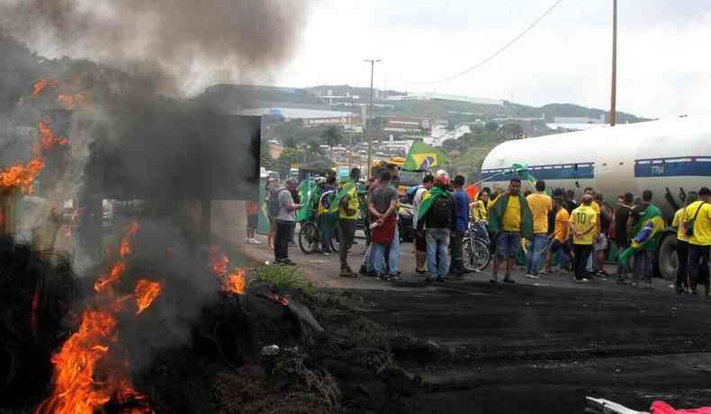 Fogo em primeiro plano, ao fundo pessoas com camisas amarelas e bandeiras do Brasil, estrada interditada