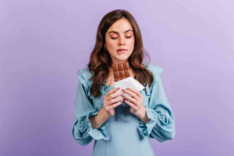 Mulher branco com cabelos castanhos longos vestindo um vestido azul sob fundo roxo segurando uma barra de chocolate