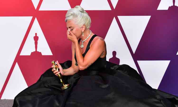 Emocionada, a cantora Lady Gaga, de joelhos e com vestido preto, est com a mo na boca e segura a estatueta do Oscar 