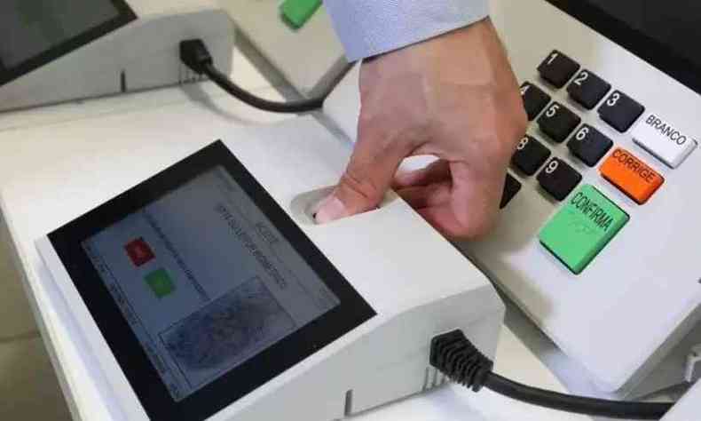 Imagem da urna eletrônica utilizada nas eleições. Há uma mão com o dedo no equipamento de biometria